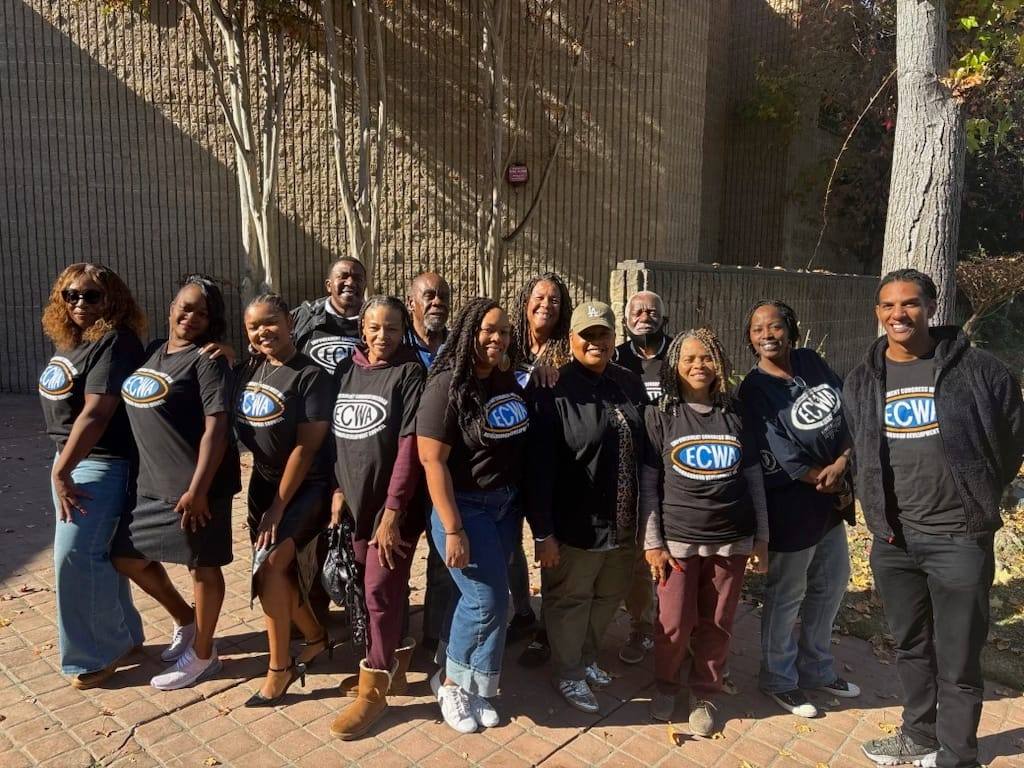 Thirteen ECWA board members and volunteers in matching black ECWA T-shirts smiling together on a brick walkway in front of a tree on a sunny day