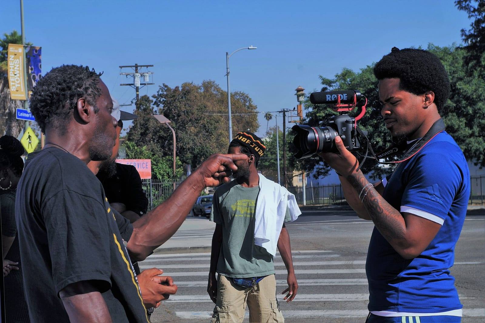 A filmmaker holds a camera while two men talk on a sunny street corner in Leimert Park, with an Art + Practice banner on a lamppost