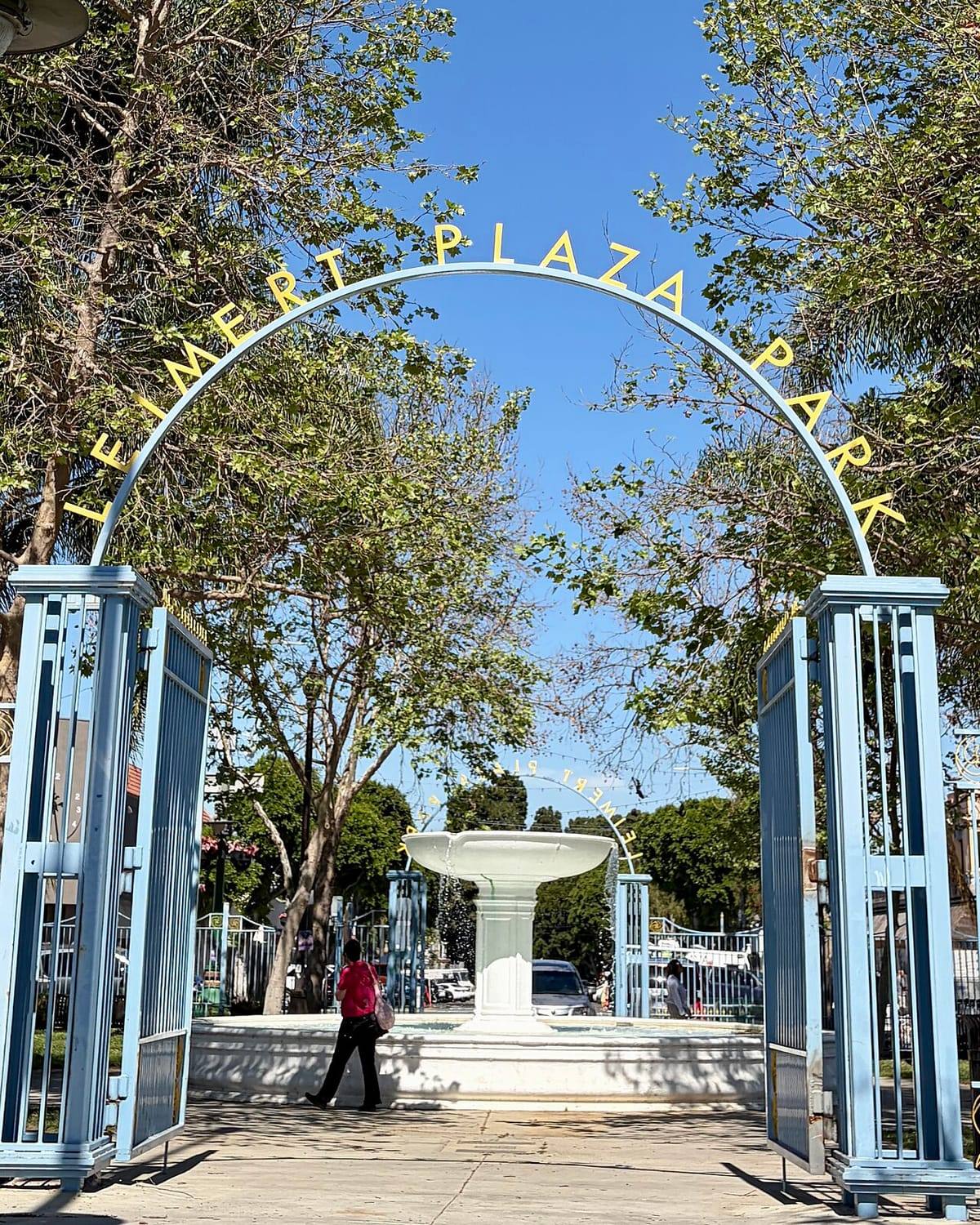 Looking through the open light-blue gates of Leimert Plaza Park, beneath a gold arch reading Leimert Plaza Park, toward the white central fountain