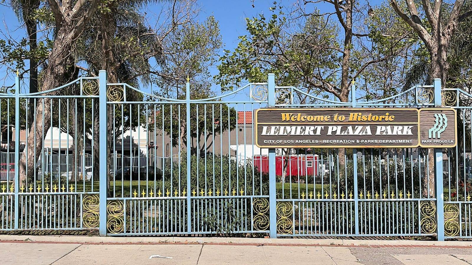 Light-blue wrought iron gates with gold scrollwork at Leimert Plaza Park, with a wooden sign reading Welcome to Historic Leimert Plaza Park