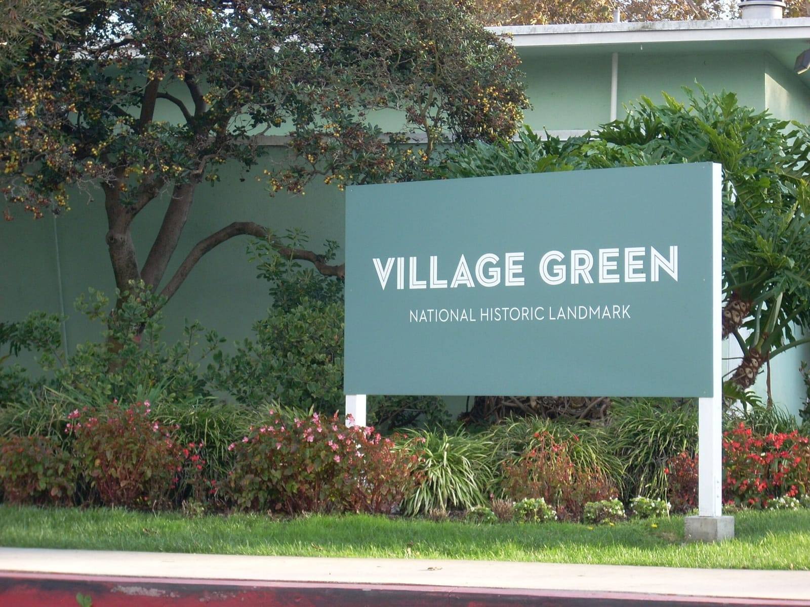A green sign reading Village Green, National Historic Landmark, set among trees and flowering shrubs